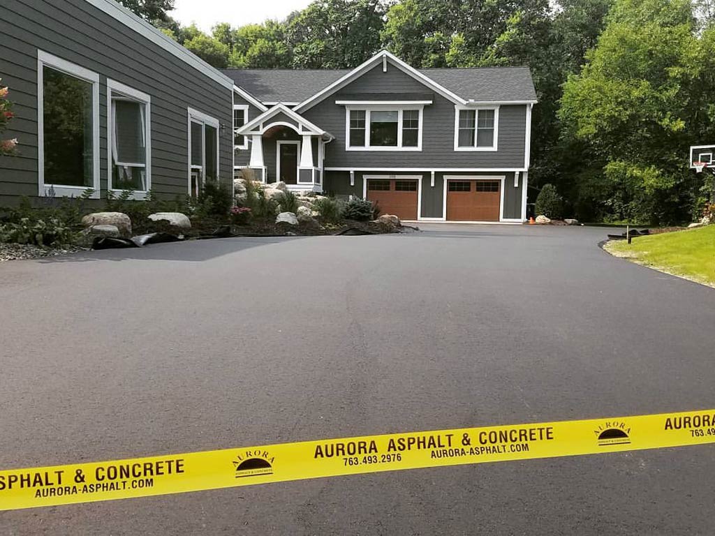 Newly paved asphalt driveway leading to a gray two-story house with a two-car garage, landscaped yard, and yellow caution tape in Minnesota.