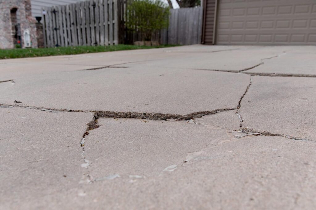 Close-up view of a cracked and sinking concrete driveway. The surface of the driveway has multiple large cracks, with visible uneven sections where parts of the concrete have sunk. The cracks are wide and uneven, indicating significant structural issues.