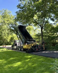 A paving project underway on a sunny day in a wooded suburban area. A large dump truck with its bed tilted is unloading asphalt into a yellow asphalt paver labeled 'Aurora.' Two workers, one in a high-visibility vest, are operating the paver while smoothing out the freshly laid asphalt on a driveway. Lush green grass and trees surround the scene, creating a peaceful backdrop for the construction activity.