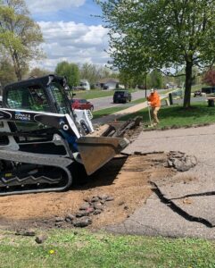 An asphalt removal project in progress. A skid steer loader is breaking and lifting crumbled asphalt from a driveway. A worker in an orange high-visibility jacket and hard hat is standing nearby, holding a shovel and preparing the site. The scene is set on a sunny day in a suburban neighborhood with green lawns, trees, and houses in the background