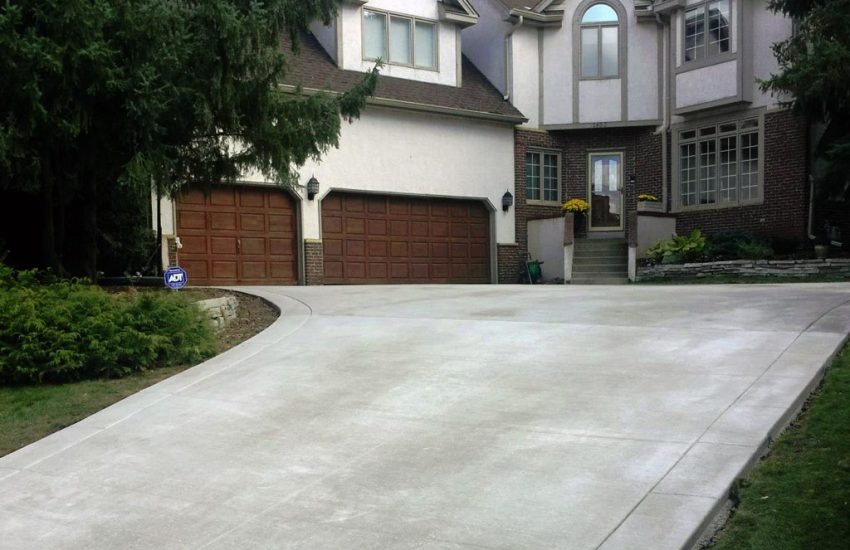 A large, smooth concrete driveway in Minnesota leading to a two-story house with brown garage doors, surrounded by green bushes and trees.